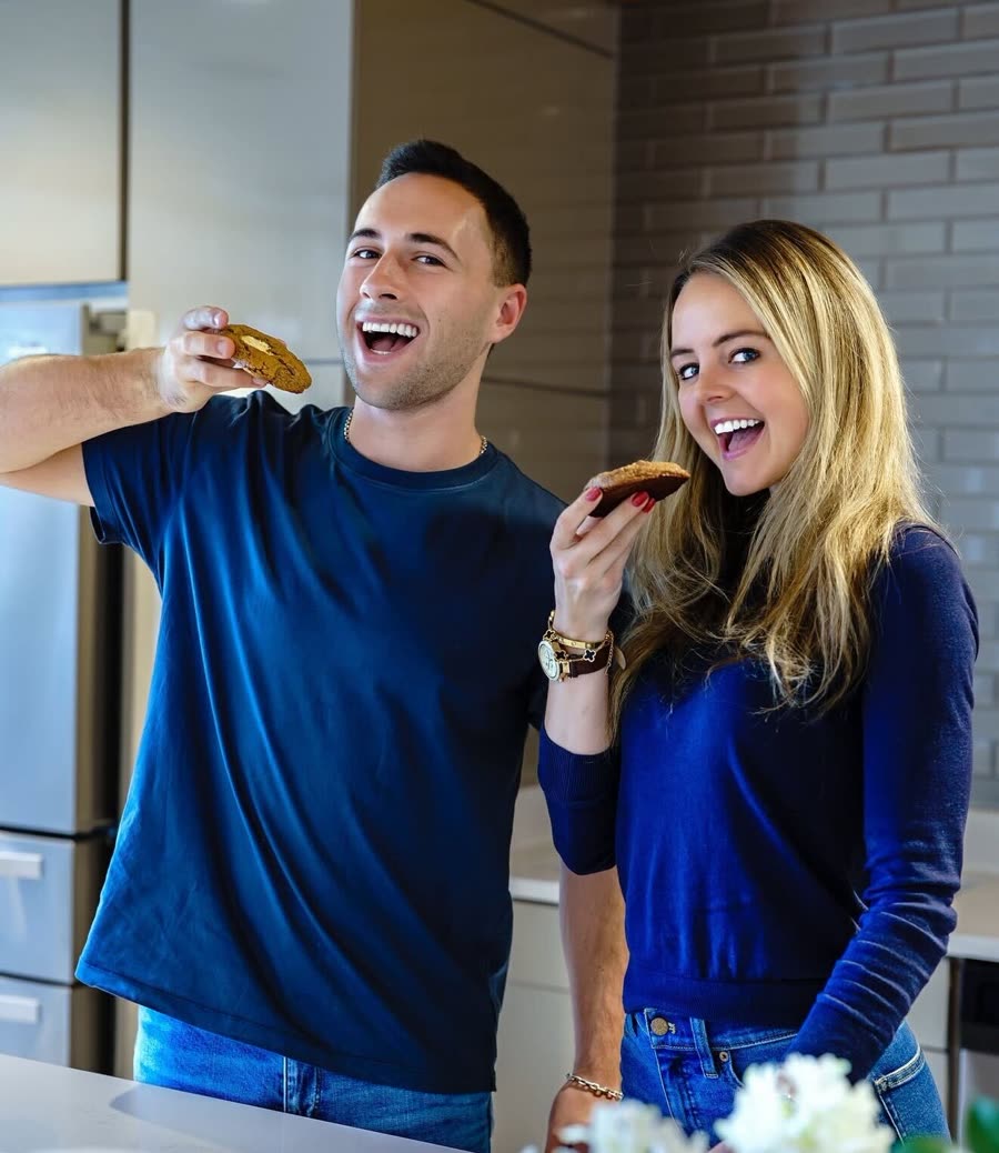 Two friends laughing in a kitchen, each holding up a Sweet Addison's cookie