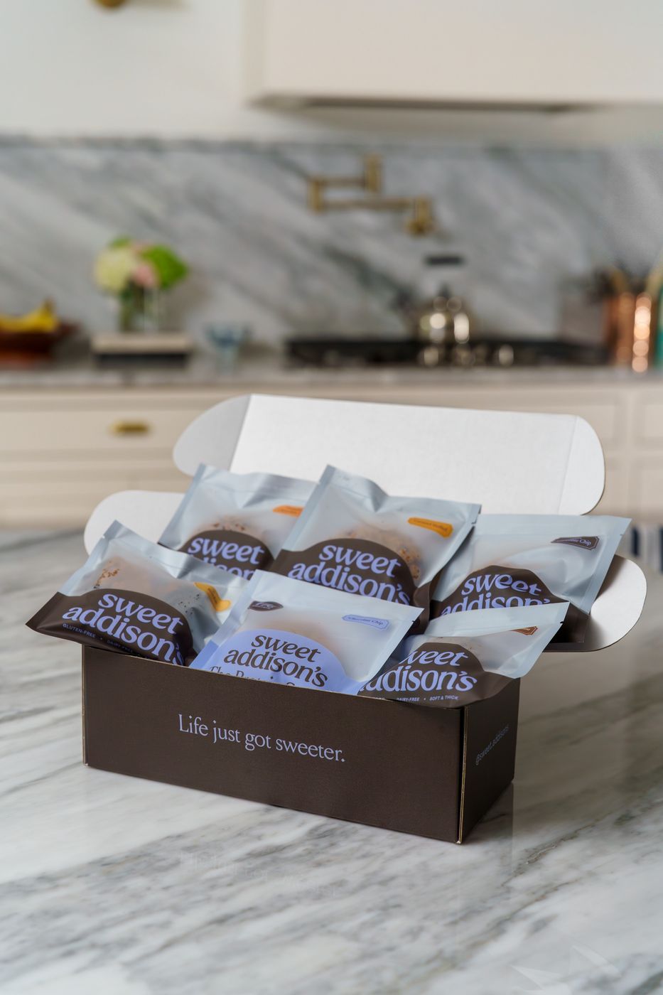 An open Sweet Addison's box of cookie pouches on a kitchen counter, home kitchen blurred behind it